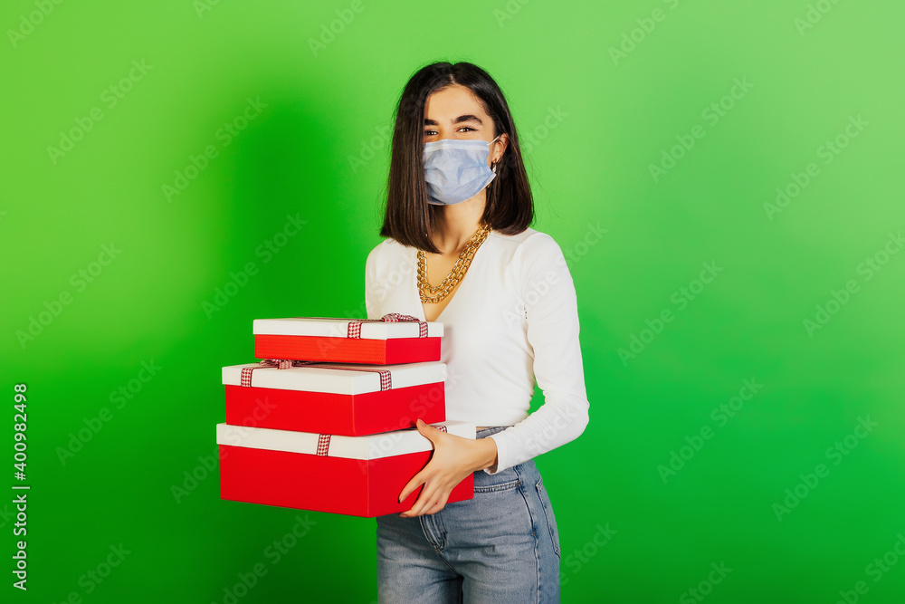 Young smiling girl with a medical mask holds gifts for the New Year or Christmas in her hands. Green studio background. Young smiling girl with a medical mask holds gifts for the New Year or Christmas in her hands. Green studio background.