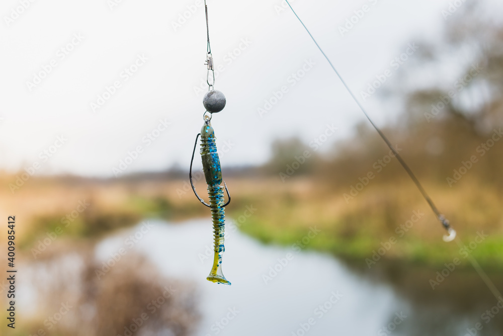 Silicone bait for fishing hanging on a hook on the background of the river. Tackle for fishing. Hobie fishing in nature Silicone bait for fishing hanging on a hook on the background of the river. Tackle for fishing. Hobie fishing in nature