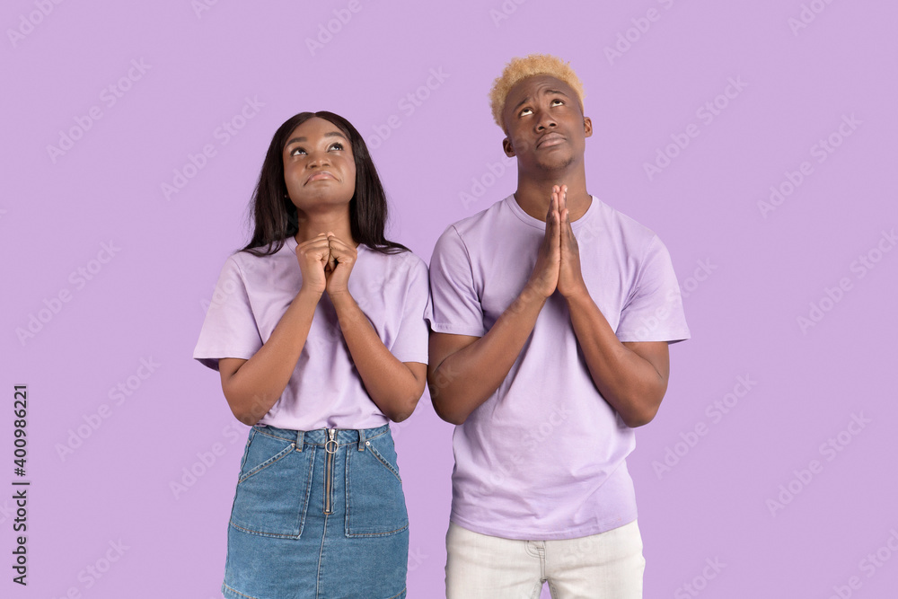 Religious black couple putting hands together in prayer, lifting their eyes up, asking God for help on lilac background Religious black couple putting hands together in prayer, lifting their eyes up, asking God for help on lilac background