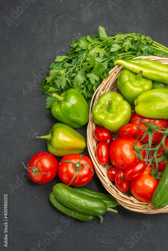 Vertical view of fresh vegetables red tomatoes with stems green peppers and cucumbers inside and outside of basket on dark background