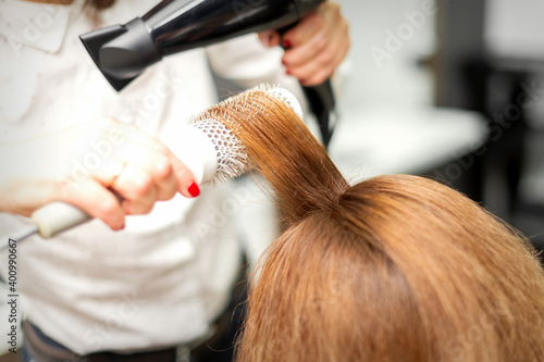 Close up of hairdresser drying long red hair with a hairdryer and round brush in a beauty salon