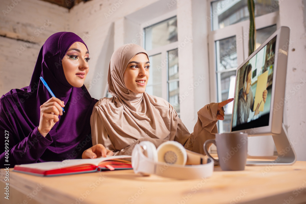 Learn. Happy and young two muslim women at home during lesson, studying near computer, online education. Culture, traditions, modern people. Watching at PC monitor, shopping or talking. Learn. Happy and young two muslim women at home during lesson, studying near computer, online education. Culture, traditions, modern people. Watching at PC monitor, shopping or talking.
