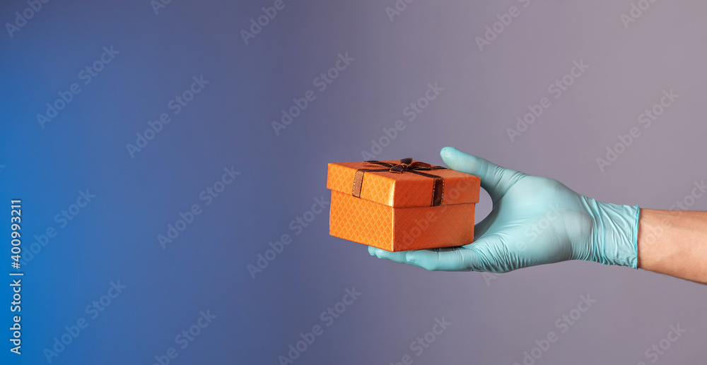 Fototapeta premium A man's hand in a blue protective glove is holding a gift box on a blue and gray background. Concept of a lifestyle in a pandemic