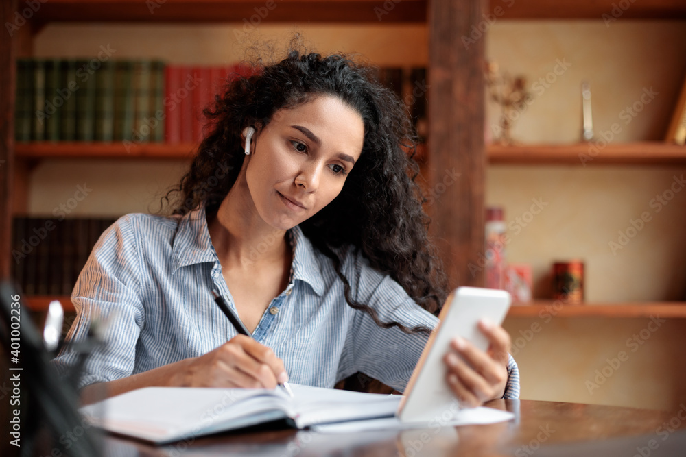 Woman sitting at desk, using smartphone and writing in notebook Woman sitting at desk, using smartphone and writing in notebook