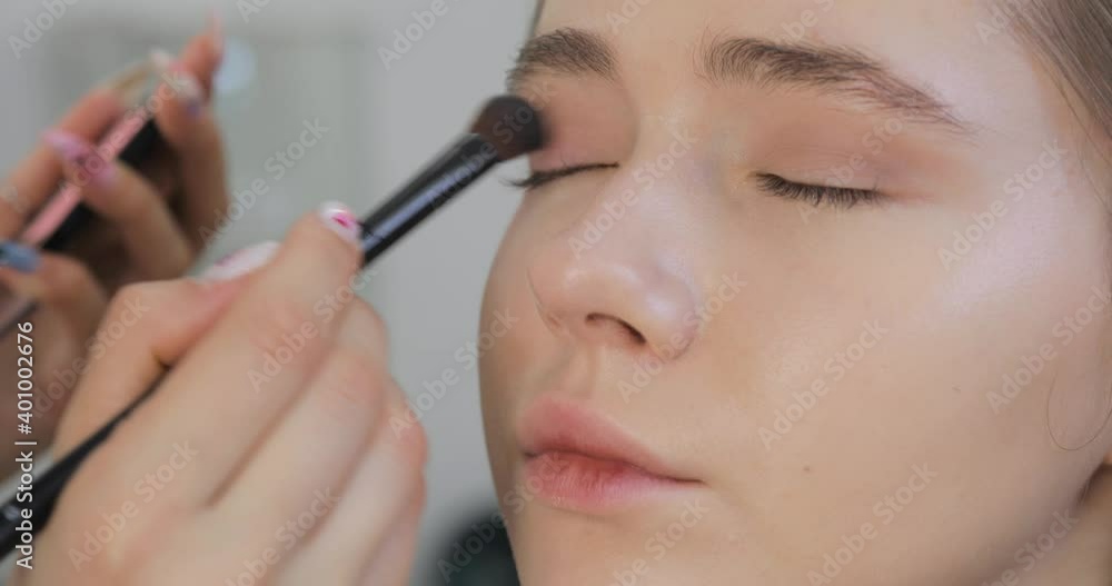 Professional makeup artist applies eye shadows to upper eyelids of model eyes using cosmetic brush, face closeup. Stylist visagist is doing makeup for young woman. Beauty industry concept. Professional makeup artist applies eye shadows to upper eyelids of model eyes using cosmetic brush, face closeup. Stylist visagist is doing makeup for young woman. Beauty industry concept.