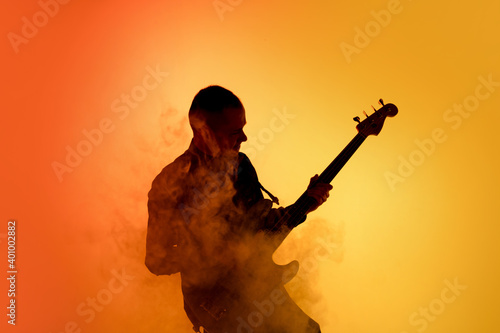 Smoke. Silhouette of young male guitarist isolated on orange gradient studio background in neon light. Beautiful shadow in action, performing. Concept of human emotions, expression, ad, music, art.