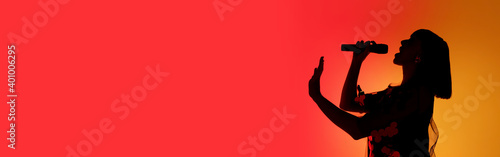 Flyer. Silhouette of young female singer isolated on orange gradient studio background in neon light. Beautiful shadow in action, performing. Concept of human emotions, expression, ad, music, art.