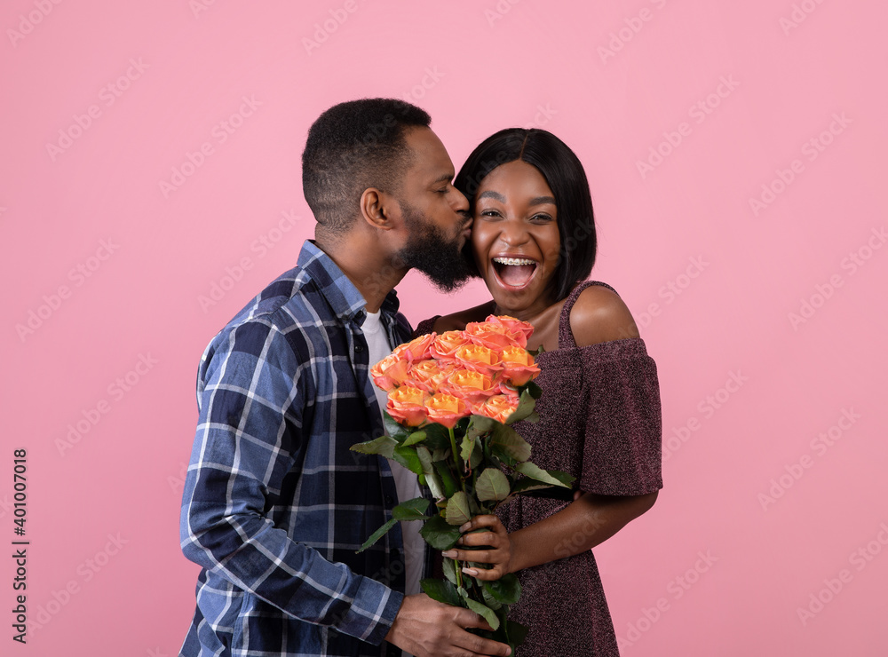 Affectionate African American guy kissing his happy girlfriend and giving her bouquet of roses on pink studio background Affectionate African American guy kissing his happy girlfriend and giving her bouquet of roses on pink studio background