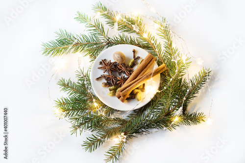 spices for mulled wine on a small white plate on a white background with green branches of a Christmas tree and a burning garland of warm yellow color. the view from the top