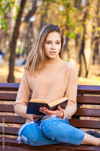 Beautiful blonde girl with a book sits on a bench in an autumn park © Andrey_Arkusha