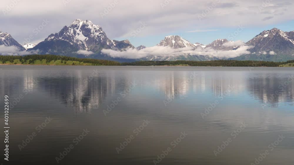 A summer morning view of a very calm Jackson Lake in Grand Teton National Park with the Tetons in the background and very low clouds drifting by A summer morning view of a very calm Jackson Lake in Grand Teton National Park with the Tetons in the background and very low clouds drifting by