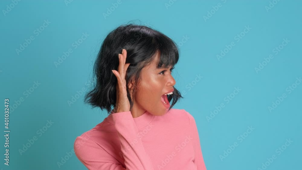 Cute curious woman keeps hand near ear tries to overhear something very interesting listens somebody secret looks away likes collecting rumors poses against blue background. Eavesdropping concept Cute curious woman keeps hand near ear tries to overhear something very interesting listens somebody secret looks away likes collecting rumors poses against blue background. Eavesdropping concept
