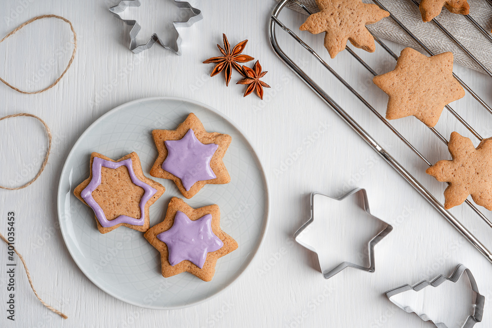 Decorating of sweet savory ginger cookies or gingerbread with violet icing on gray plate cooked for christmas winter holidays on white wooden table with cutters, anise and grate. Horizontal, flat lay Decorating of sweet savory ginger cookies or gingerbread with violet icing on gray plate cooked for christmas winter holidays on white wooden table with cutters, anise and grate. Horizontal, flat lay