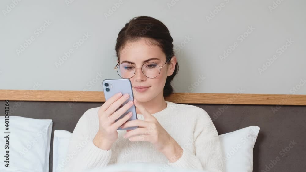 Young pretty girl lies in bed and using mobile phone indoors at home Young pretty girl lies in bed and using mobile phone indoors at home