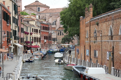 a venetian canal full of boats  with tenement houses near the water s edge  buildings of various colors  pavements with people walking