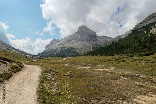 A gravelled road leading through Italian Dolomites. High, sharp mountains around. The slopes and meadow are green, higher parts barren and stony. Overcast. Remote and desolate place. Freedom
