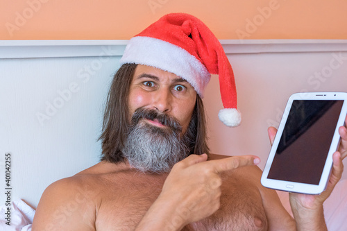Close-up view of bearded with long hairs caucasian man in Santa Claus hat uses a tablet PC laying on the bed. He looks at the camera in surprise, pointing at something on the tablet. Holiday, festive photo