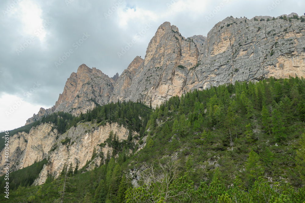 A view on a sharp and stony mountain wall in Italian Dolomites. The massive mountain is surrounded by clouds. Lower parts overgrown with trees. Remote and desolate landscape. Silence before the storm