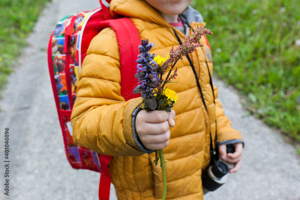 niño con cazadora de invierno amarilla y mochila roja sujeta con la mano un puñado de flores silvestres