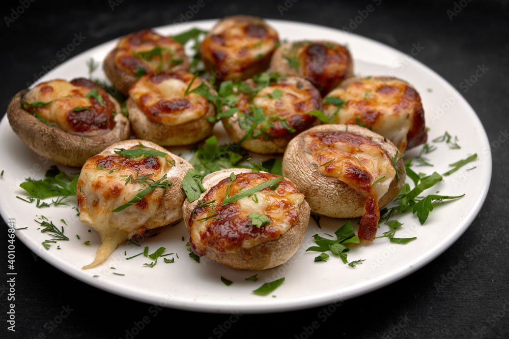 Baked stuffed mushrooms champignons, with cheese and herbs, on a white plate, on a black background