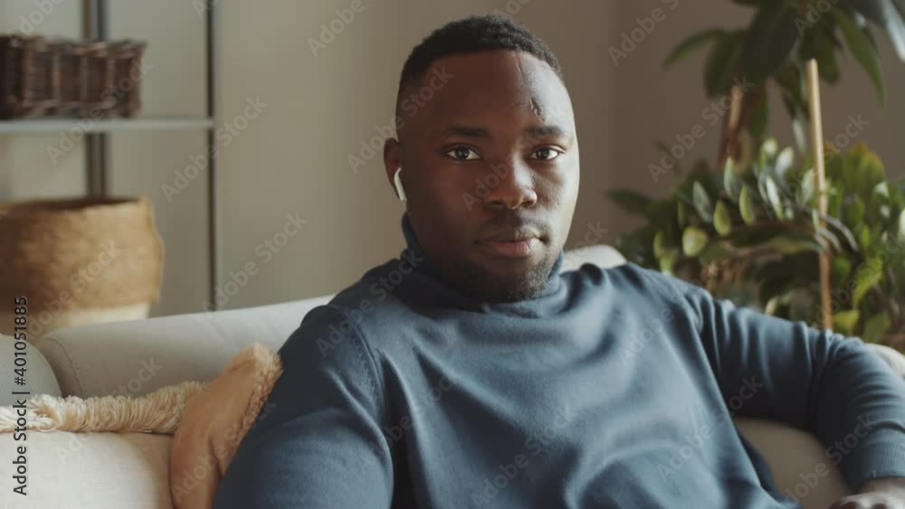 Tilt up shot of young Afro-American man in wireless earphones using laptop and then looking at camera while sitting on sofa at home Tilt up shot of young Afro-American man in wireless earphones using laptop and then looking at camera while sitting on sofa at home