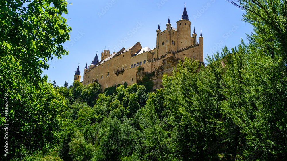 Castle of Segovia on top of the mountain with green vegetation and blue sky.
