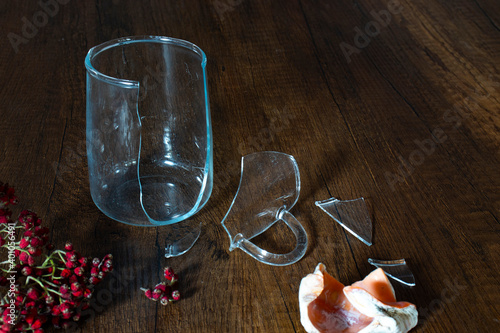 Broken glass cup and seashell are isolated on wooden background.
