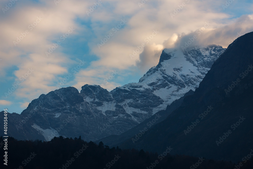 Snow-capped peaks in clouds and blue skies. Winter mountains, alpine skiing. Sunny snowy landscape. Panoramic sunset mountain view. Rocks and white slopes covered with glaciers