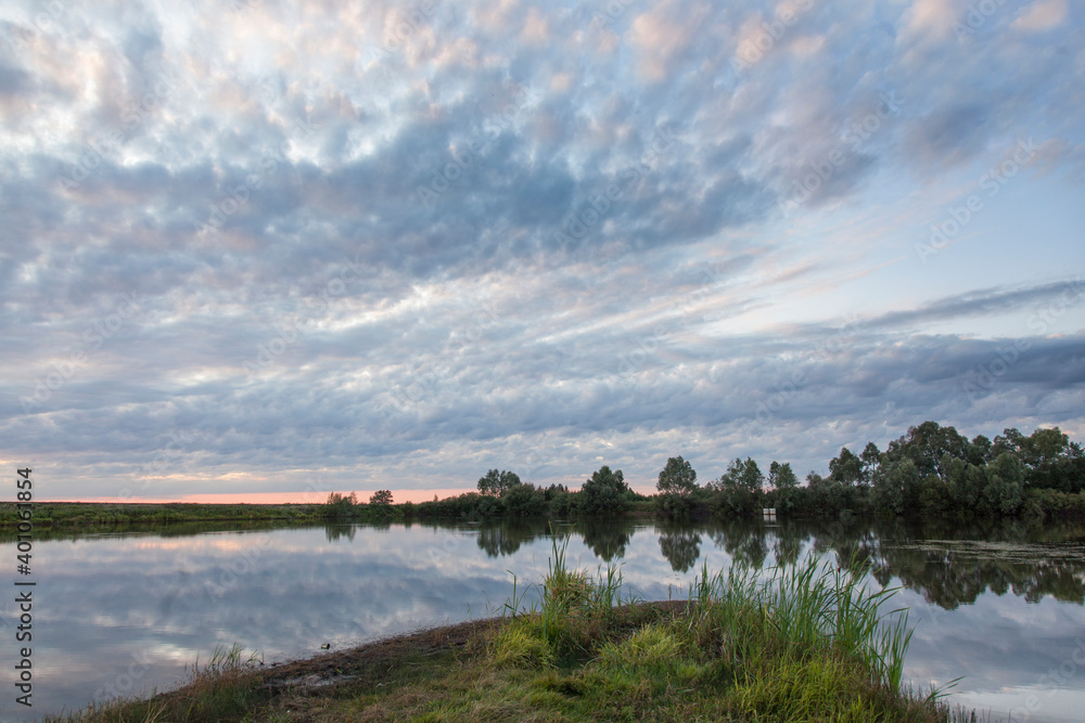 Obraz premium Autumn evening on the lake, the banks of which are overgrown with grass and reeds. In the sky, the clouds are colored by the light of the setting sun.