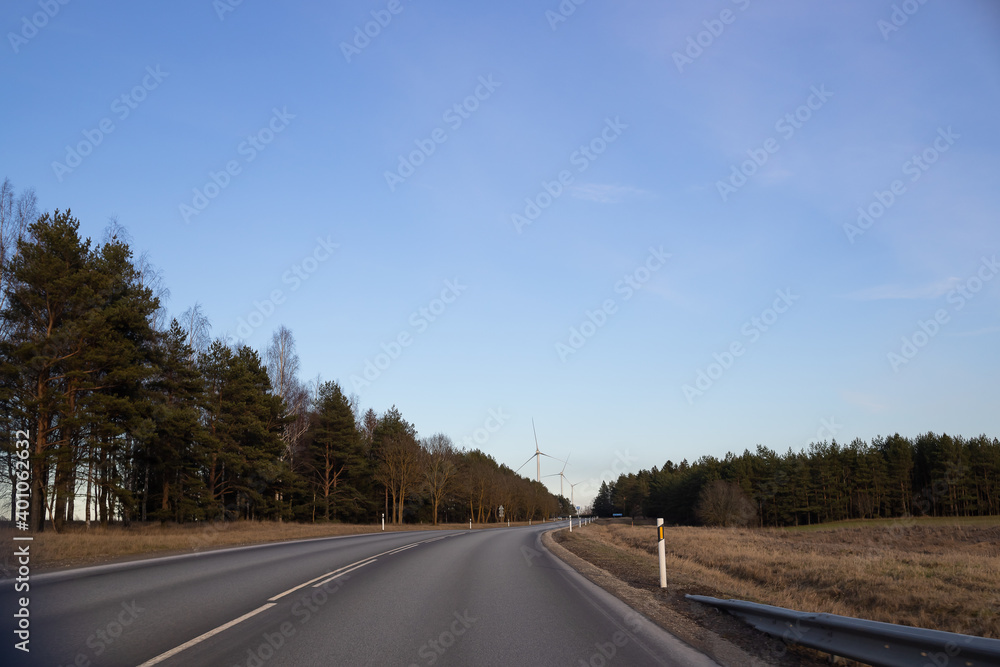 The road goes far beyond the forest and windmills.Wind turbine. Empty road in foreground, blue sky with clouds. Alternative energy source, production and power generation. Ecology and freedom concept.