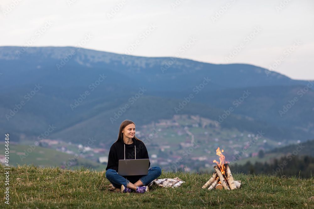 young female freelancer working on laptop in the mountains in the evening. Tourist girl sitting near campfire and having fun. Copy space.