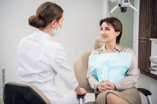 A portrait of a woman with a toothy smile sitting on the dental chair and talking to her doctor.