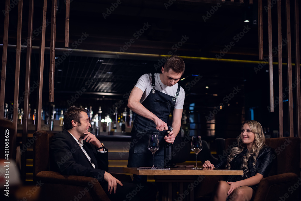a young bartender opens a bottle of red wine for a man and a woman at a table in a restaurant.