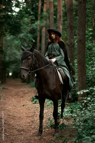 Young beautiful woman in a suit, green dress, corset, black topm hat on a black horse on nature in the forest. fairy tale, creative photo session of a girl with a horse © Владимир Николаев