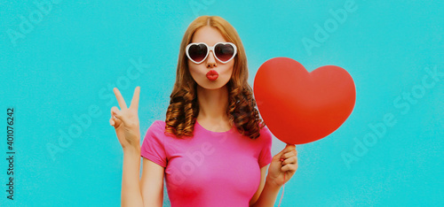 Portrait of happy young woman holding red heart shaped balloon and blowing lips sending sweet air kiss on a blue background