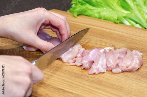 Woman in an apron in the kitchen slicing chicken fillet on an oak board. Vegetables in the kitchen