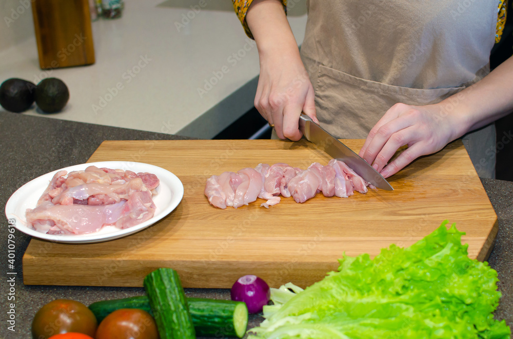 Woman's hands are cutting chicken fillet on a cutting board. The chef prepares chicken in the kitchen.