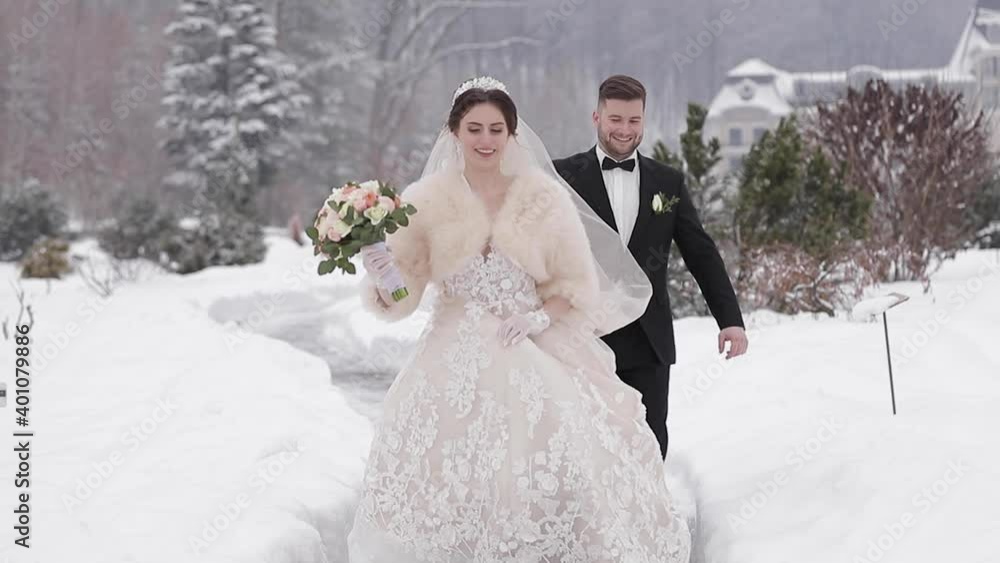 Elegant bride in white fur coat stand with handsome bearded groom in front of snow covered pine trees Elegant bride in white fur coat stand with handsome bearded groom in front of snow covered pine trees