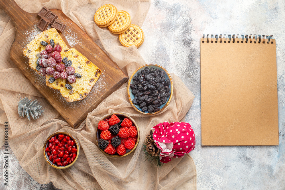 top view raisin cake on cutting board bowl with pomegranate raspberry candies and raisins biscuits on beige tablecloth a notebook on marble ground