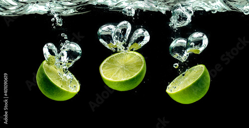 Bunch of lime fruits halves splashing with bubbles into water isolated against black background. Citrus theme, panorama
