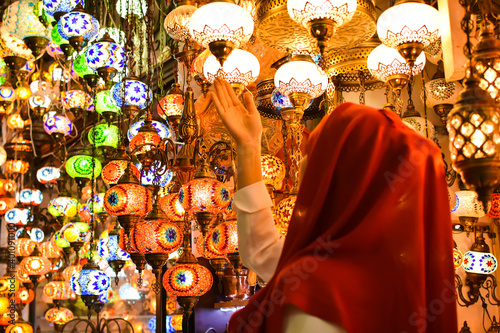 Young pretty tourist Muslim woman with red scarf looking on Traditional Turkish lamps in souvenir shop. Woman in hijab shopping in the Grand Bazar. Shopping tour. photo