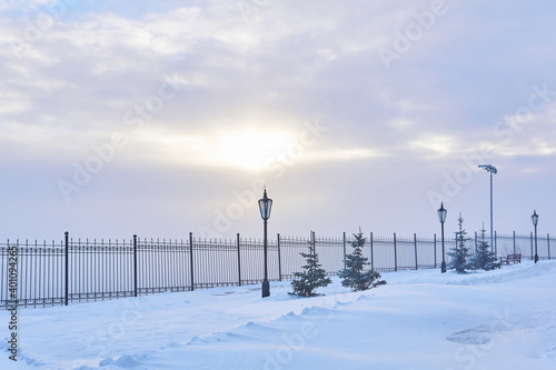 winter snowy park with a fence, lanterns and sunlight shining through the clouds