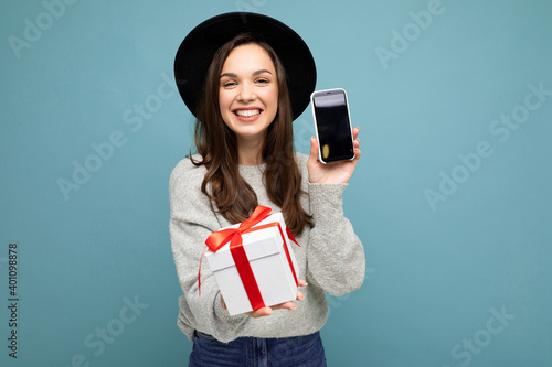 Shot of pretty smiling positive young brunet woman isolated on blue background wall wearing black hat and grey sweater holding gift box showing smartphone screen display for mockup and looking at photo