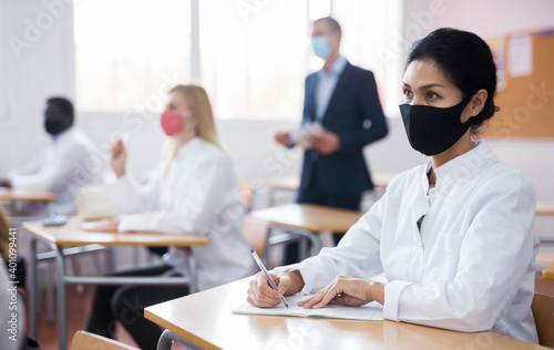 Tableau sur toile Adult asian female medical student in protective face mask studying in classroom with group, writing lectures in workbook