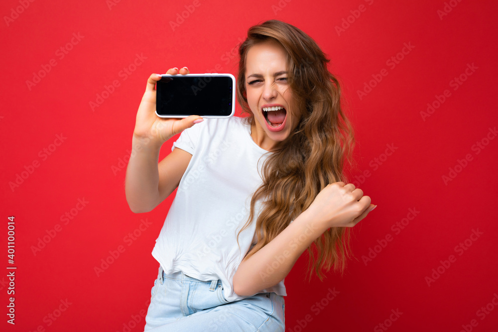 Photo of beautiful overjoyed young blonde woman good looking wearing white t-shirt standing isolated on red background with copy space holding phone showing smartphone in hand with empty screen Photo of beautiful overjoyed young blonde woman good looking wearing white t-shirt standing isolated on red background with copy space holding phone showing smartphone in hand with empty screen