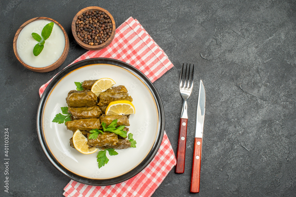 top view stuffed grape leaves on white plate bowls of natural yogurt and black pepper knife and fork napkin on dark background with copy space top view stuffed grape leaves on white plate bowls of natural yogurt and black pepper knife and fork napkin on dark background with copy space