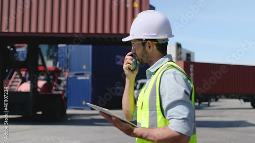 Foreman or cargo container worker walk along and in front of crane with container tank and also use walkie talkie to communicate with co-worker in workplace area.