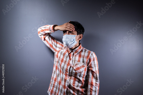 Depressed and sick young woman in protective mask holding her head on gray background.