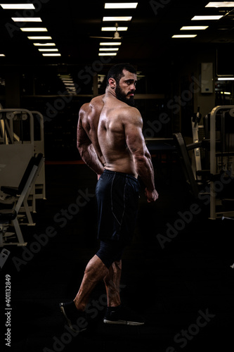 strong young bearded caucasian man with sport physique body standing in dark fitness gym showing powerful back muscle