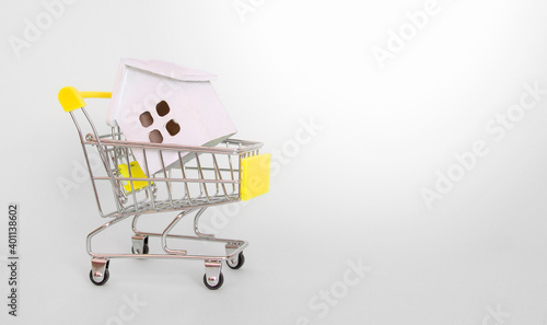A white wooden house lies in a grocery cart with a yellow handle on a gray background. Side view. Horizontal format. Copyspace